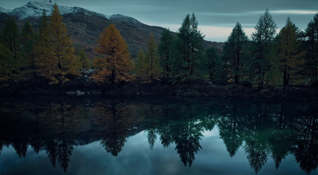 A still lake at dusk reflecting autumn trees, with a break of light on the far horizon
